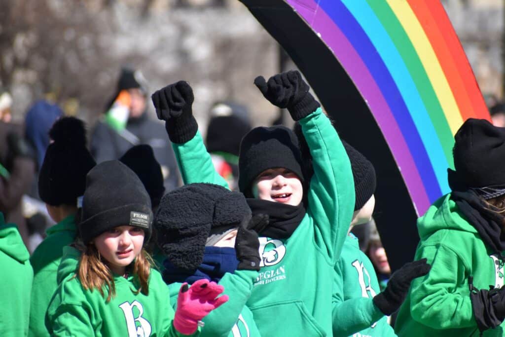 Children in green sweatshirts cheering under a rainbow balloon arch during Milwaukee’s St. Patrick’s Day parade.