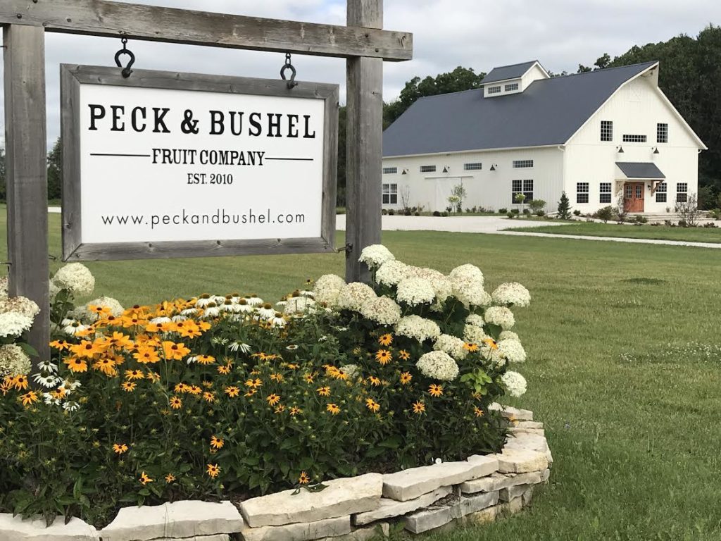 Front view of Peck & Bushel Fruit Company with their business sign and fall flowers and white barn in the background.