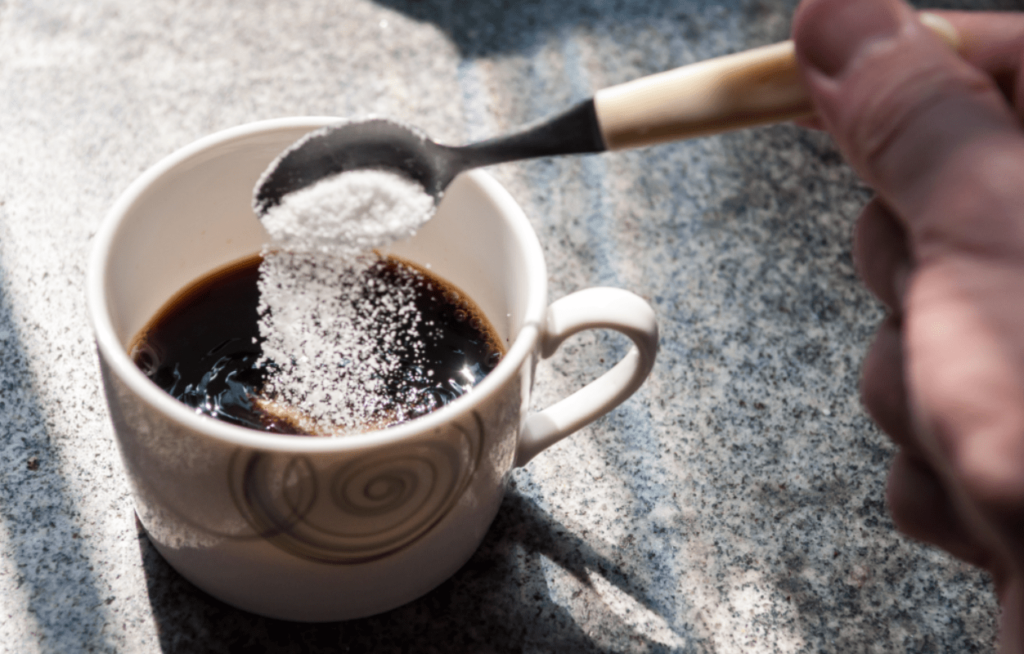 A person puts sugar in their mug of coffee.