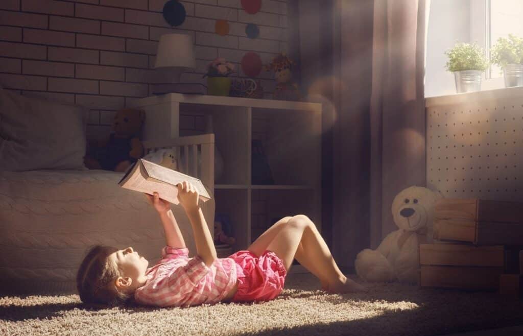 Child reading book with teddy bear and sunshine coming through the window.