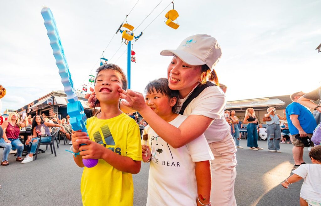 Family poses for a picture at the State Fair with colorful sky gliders in the background, and one daughter holding food on a stick.