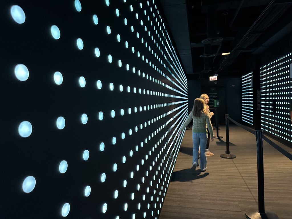 wo children walk through a neon-lit hallway with glowing dots at 360 Chicago before taking the elevator to the observation deck.