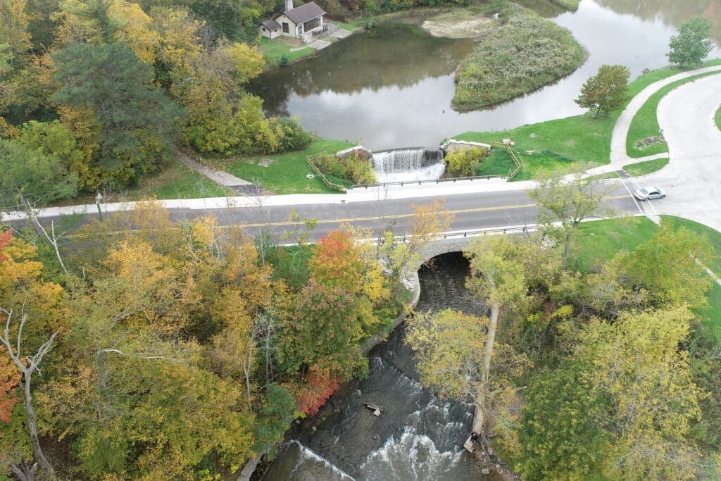 Drone view of a waterfall flowing under a road bridge near a historic building and a scenic pond in Grant Park.
