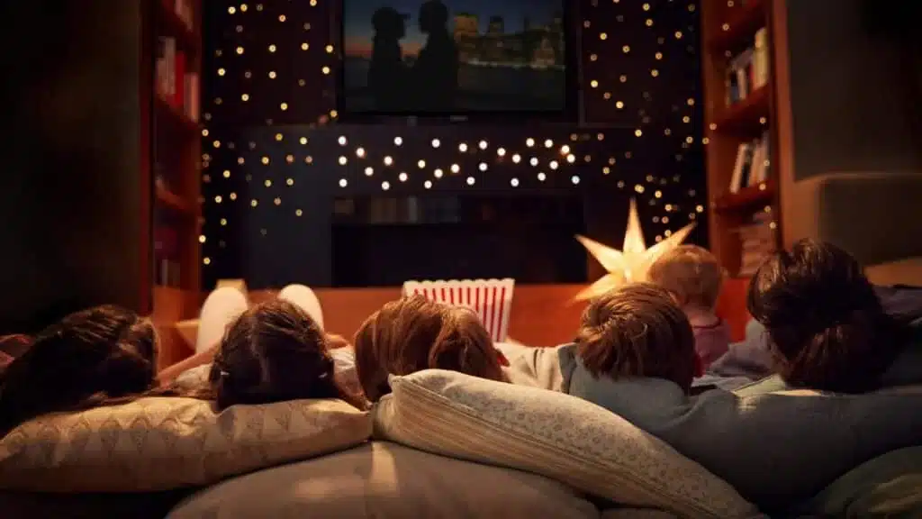Kids lying on pillows watching a movie with popcorn and string lights