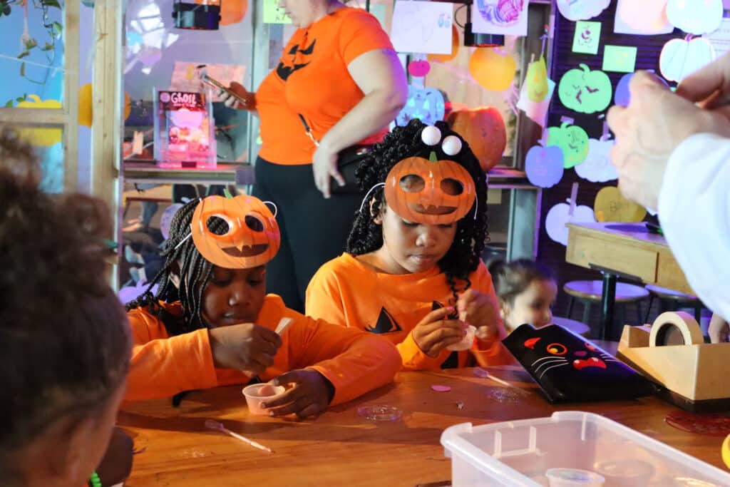 Two kids in orange pumpkin costumes work on a Halloween craft at Betty Boo's Spooktacular event at Betty Brinn Children's Museum