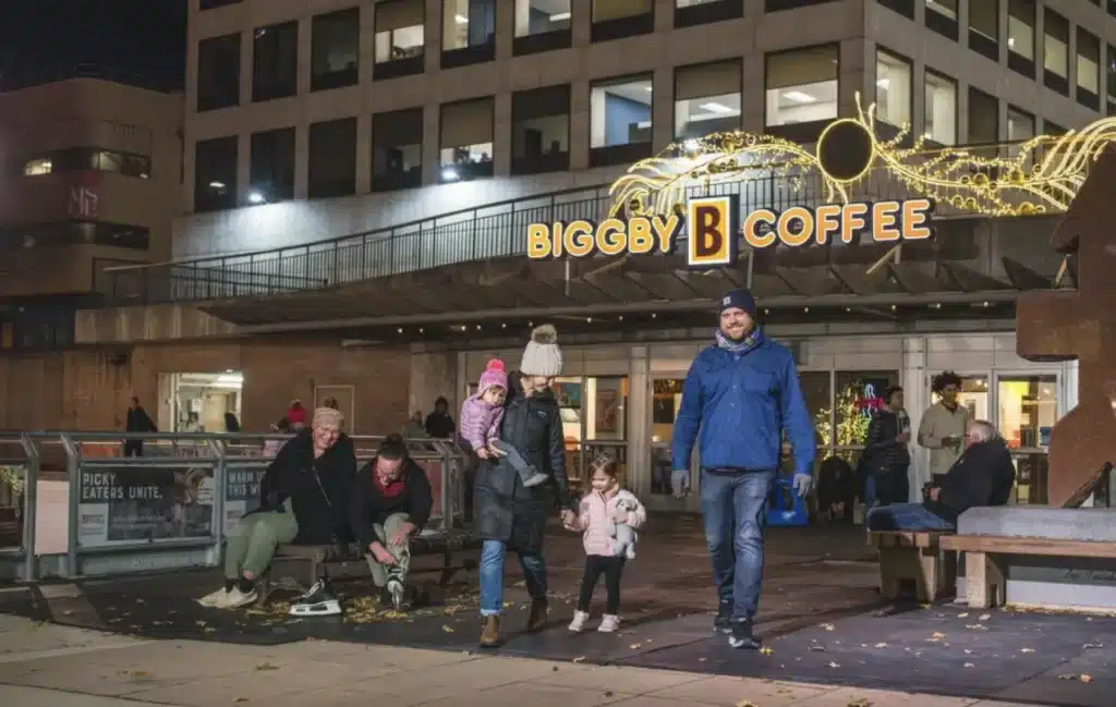 A family walks through Red Arrow Park in front of Biggby Coffee bundled up in warm clothing while another family laces their ice skates.