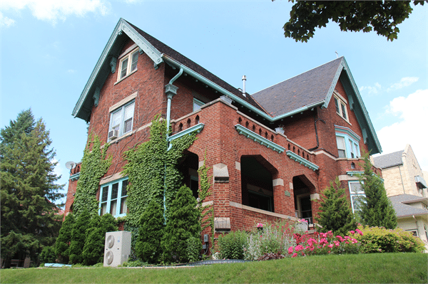 Historic red-brick Brumder Mansion in Milwaukee, covered with ivy and surrounded by colorful flowers and greenery, with gabled rooflines and teal trim under a bright blue sky.
