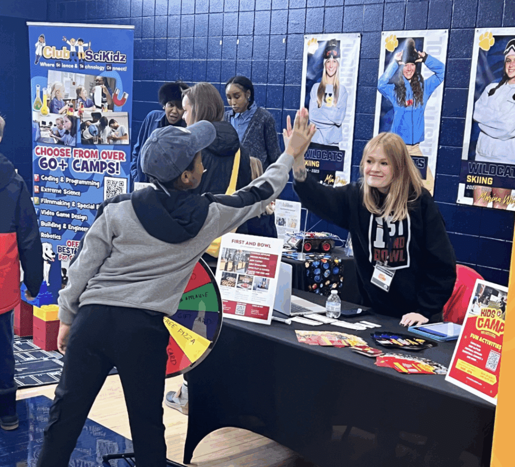 Camp fair exhibitor gives a high five to a child at the Milwaukee Camp Fair, surrounded by colorful displays and camp information.