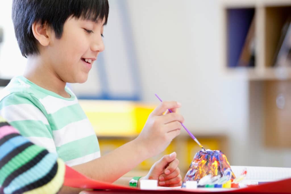 A boy painting a mini volcano science project in yellow, purple and green colors