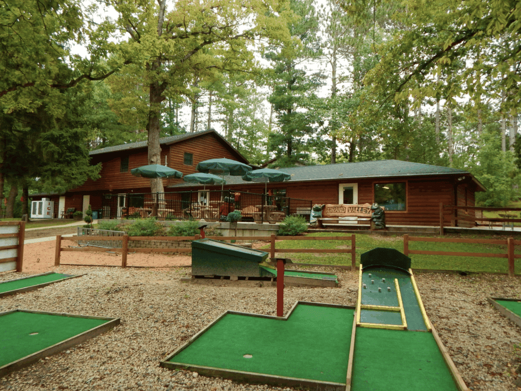 Mini golf course in front of a rustic log cabin-style building surrounded by tall pine trees at Grand Valley Campground in Dalton, Wisconsin.