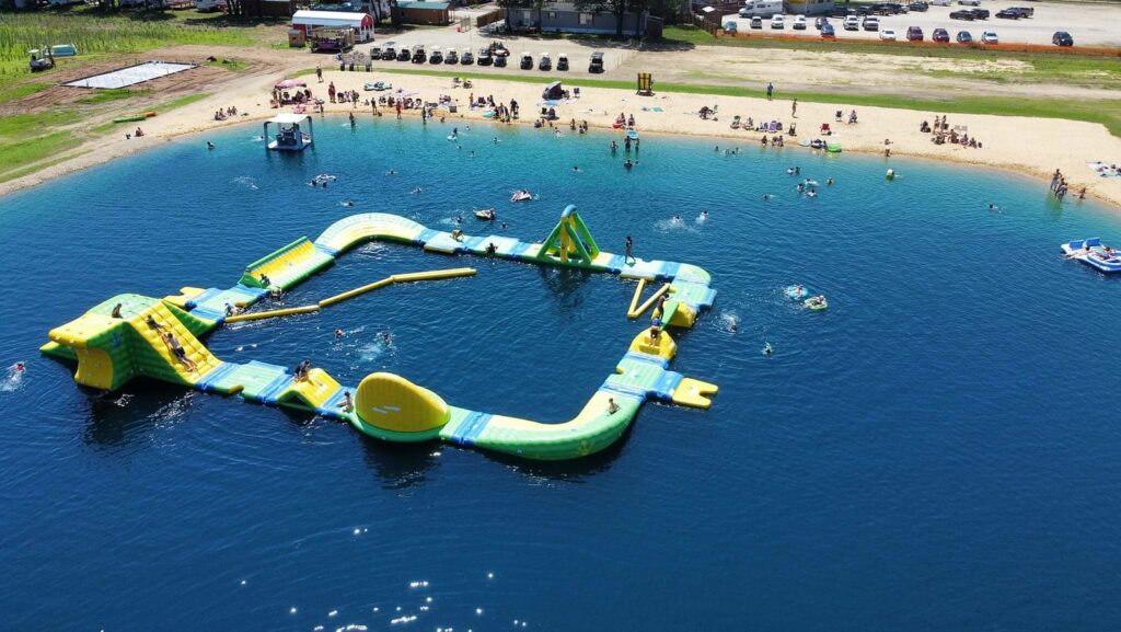 A large inflatable obstacle course and slides in bright yellow and green on clear blue water, with people swimming and relaxing on the sandy beach nearby at a family campground in Wisconsin.