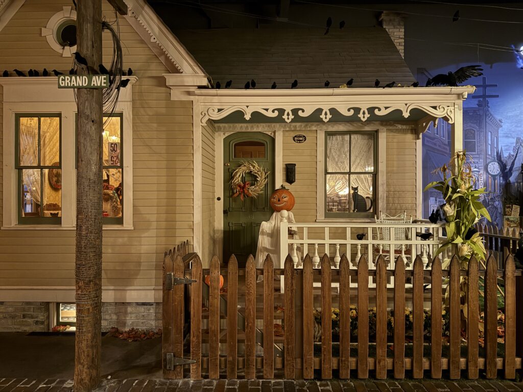 A historic-style house decorated for Halloween inside the Milwaukee Public Museum’s Streets of Old Milwaukee exhibit. A pumpkin-headed figure stands on the porch, cornstalks are tied near the fence, and dozens of black crows perch along the roofline. A black cat silhouette sits in the window, adding to the eerie vintage scene.
