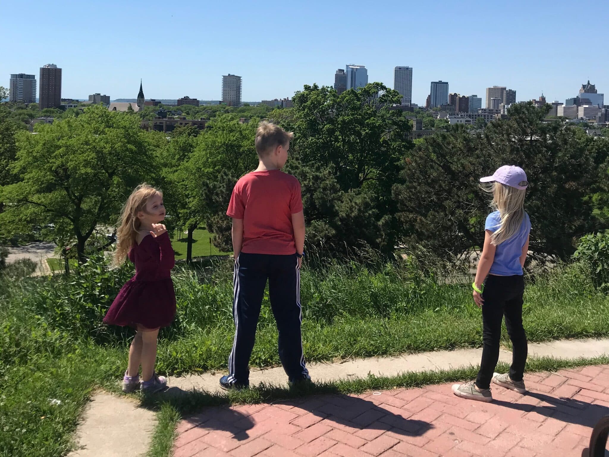 Three children standing on a hilltop, overlooking a city skyline on a bright, sunny day at Alice Bertschy Kadish Park