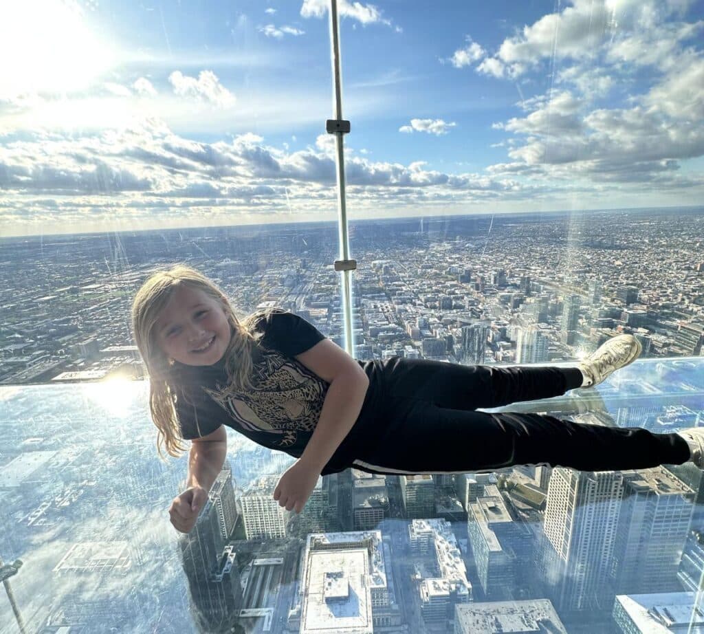 A child smiles while lying on the glass floor of The Ledge at Skydeck Chicago, looking down over the buildings below.