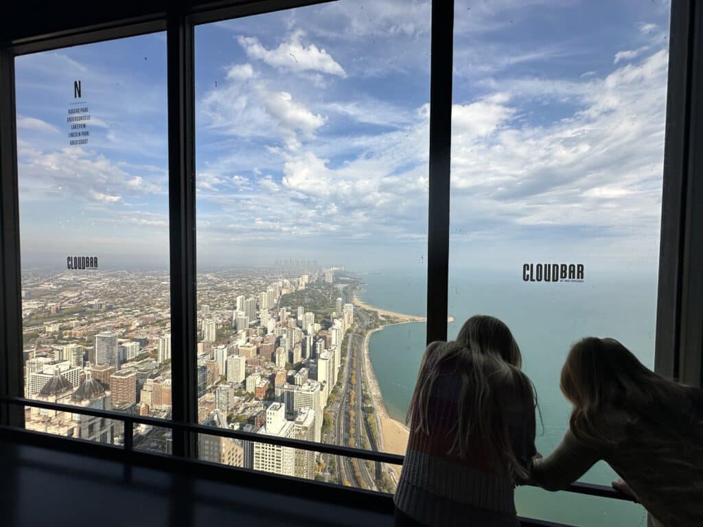 Two kids look out the large windows at 360 Chicago, spotting neighborhoods along Lake Michigan from high above the city.