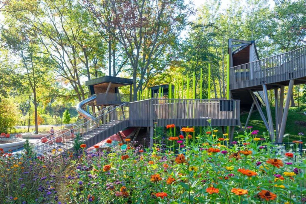 Modern wooden playground structure with slides and bridges surrounded by vibrant wildflowers and tall trees in a botanical garden setting.