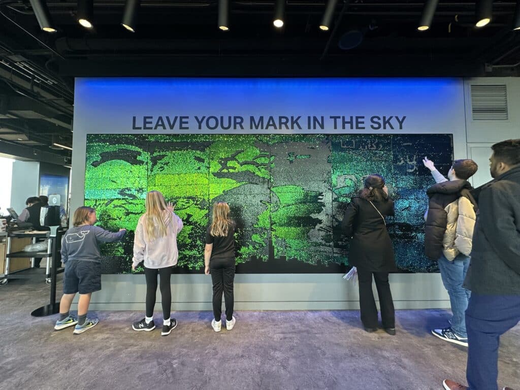 Kids interact with a sparkly green sequin wall that says “Leave Your Mark in the Sky” at Skydeck Chicago.