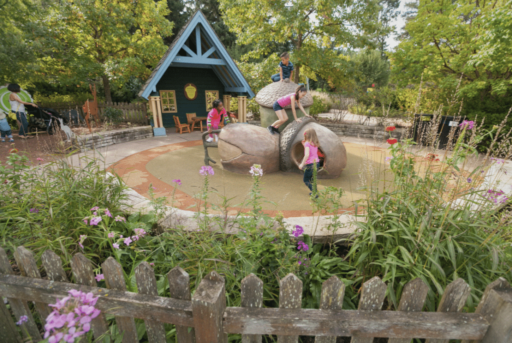 Children playing on oversized acorn sculptures in a natural playground area, with a cozy cabin-like playhouse in the background and flowers along a picket fence.