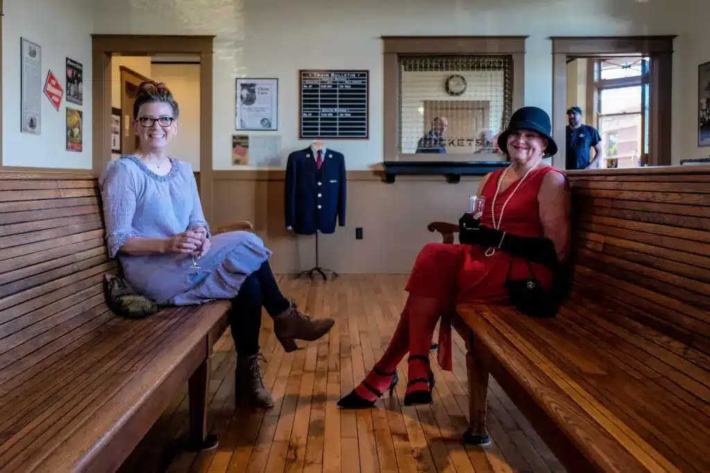 Two ladies in Roaring 20s costumes sitting across each other and smiling at the camera