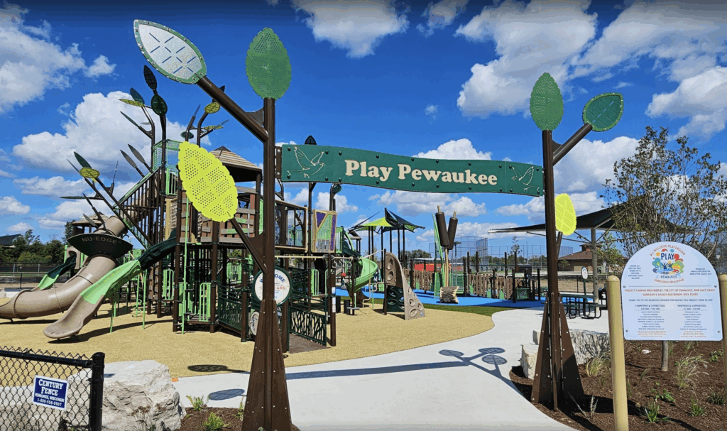Entrance to Play Pewaukee, an inclusive Lake Country–themed playground at the Pewaukee Sports Complex, featuring tree-inspired play structures, tall slides, climbing features, and a welcoming sign surrounded by bright green leaf details under a blue sky.