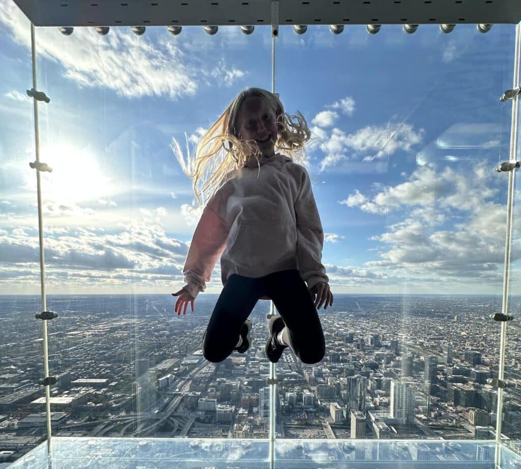 A child jumps joyfully inside The Ledge glass box at Skydeck Chicago with the city skyline visible beneath their feet.