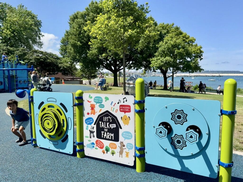 A young boy plays near the interactive play structure at South Shore Park that reads "Let's Talk The Farm" with Lake Michigan in the background on a sunny day.