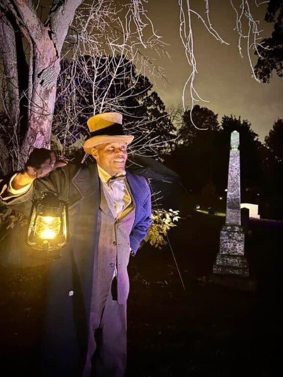 A man in an old-fashioned suit and coat holds up a lantern in the Forest Home cemetery at night during a Spirits of the Silent City tour.
