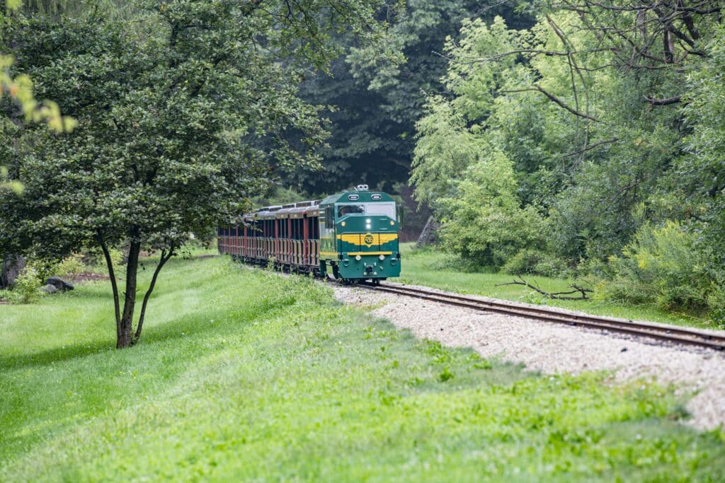 The Milwaukee County Zoo’s new Engine No. 2025, a green and yellow Safari Train locomotive, pulls passenger cars along a wooded track surrounded by lush greenery.