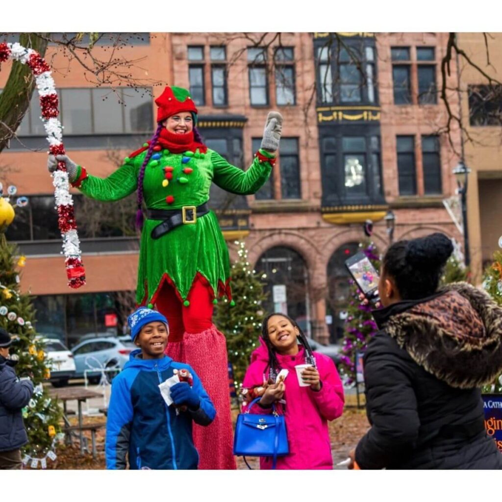 A costumed performer dressed as a festive elf stands high on stilts, smiling and holding a large decorative candy cane in an outdoor holiday setting. In front of the performer, two children—one in a blue jacket and one in a bright pink coat—smile and pose while someone nearby takes their photo. Decorated Christmas trees and buildings line the background. Cocoa With the Clauses event in Cahtedral Square Park, Milwaukee, Wisconsin.