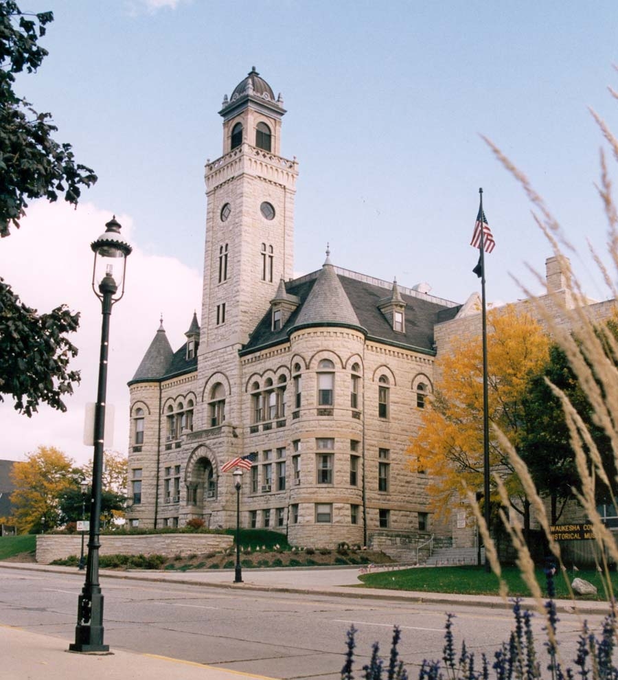 Waukesha County Historical Museum – A grand historic stone building with a clock tower, arched windows, and American flags, set against a bright sky with autumn foliage.