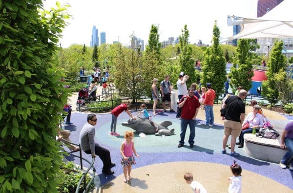Crowded playground scene with families enjoying outdoor play structures and water features, with city skyscrapers visible in the background.