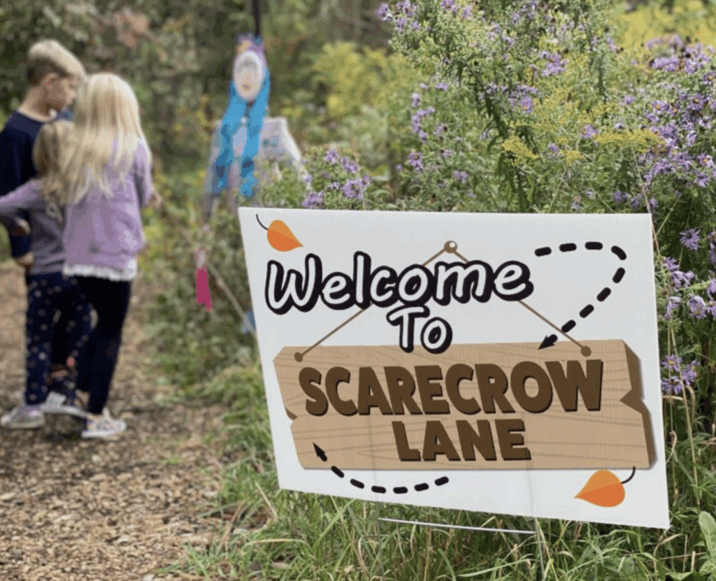 Children walking along a wooded trail at Scarecrow Lane, passing a colorful scarecrow display with a welcome sign surrounded by purple wildflowers.