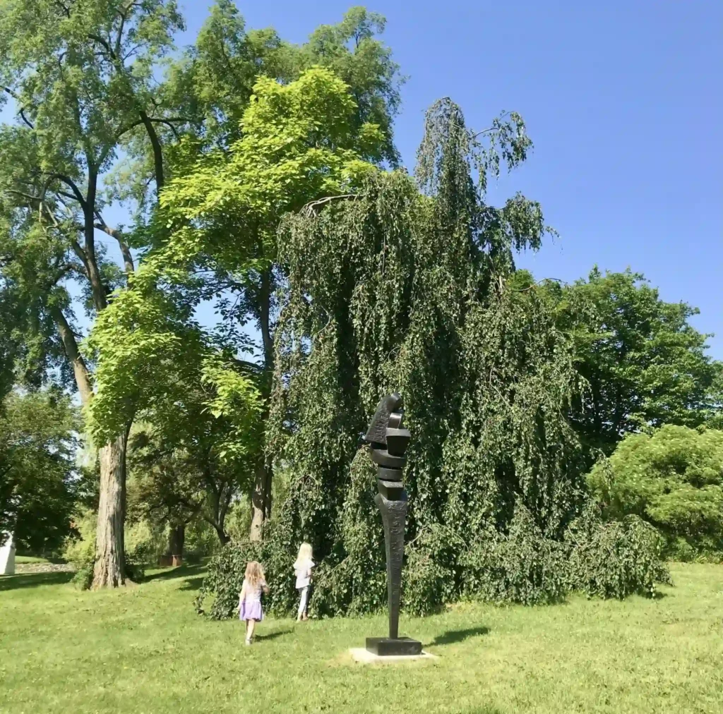 Children exploring a park near a large tree and outdoor sculpture.