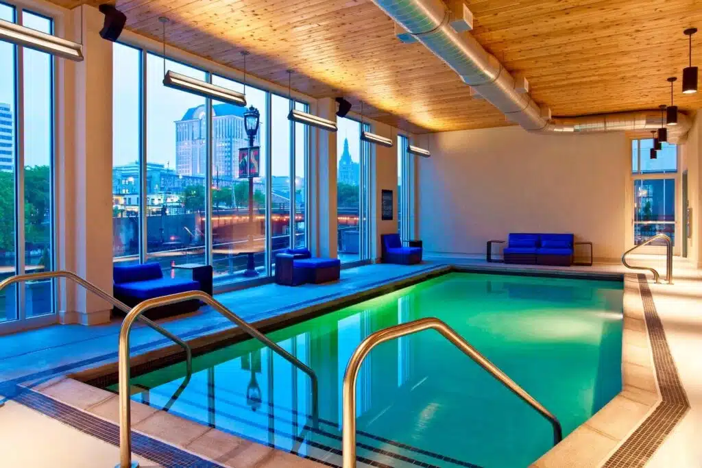 An indoor pool with wide windows and blue couches and overlooking a river and tall building