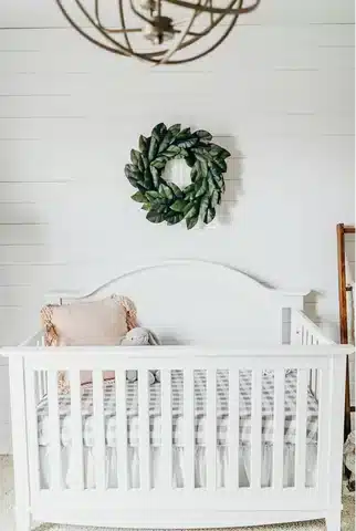 Baby girl nursery with crib with vintage style mattress, coral accent pillow and green wreath inspired by the farmhouse design.