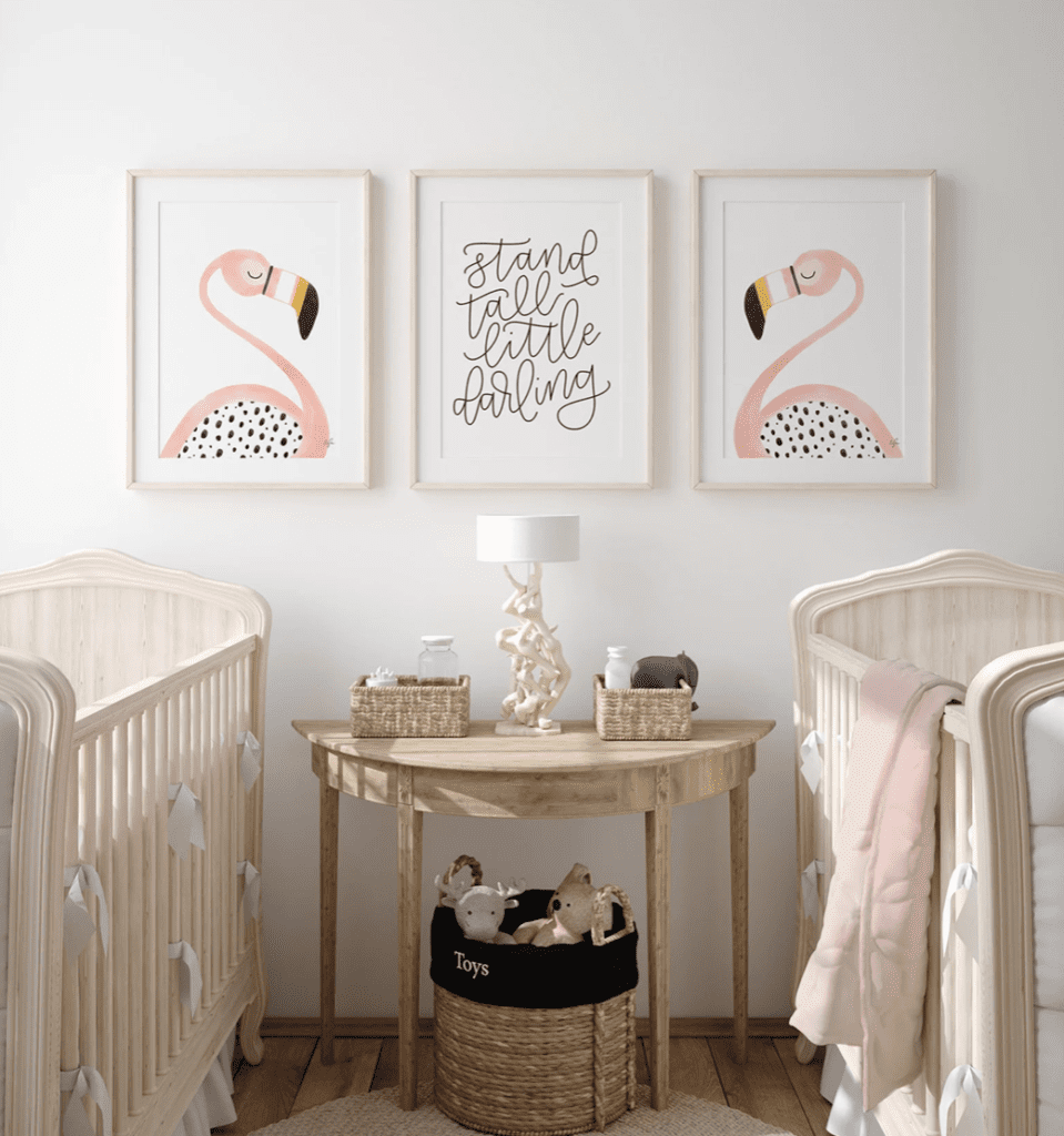Baby girl nursery with twin crib, wood console table and flamingo artwork on the wall to show flamingo themed design.