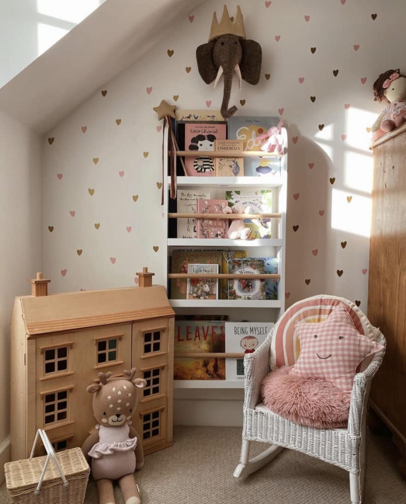 Baby girl nursery with plush elaphant wall decor, dollhouse, wicker chair, bookshelf and wallpaper with heart prints to highlight the heart theme.