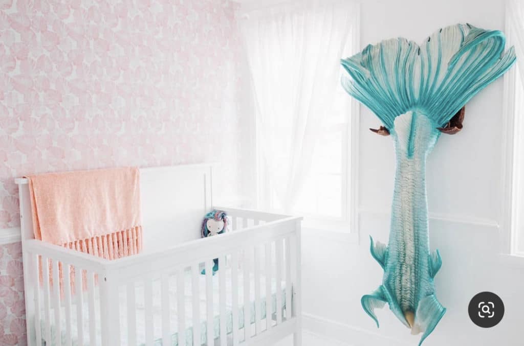 Baby girl nursery with sheer curtains, mermaid wall print and soft pink accent wall representing a mermaid inspired baby room.