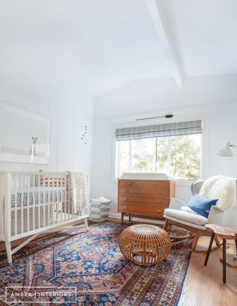 Baby girl nursery with wooden cabinets, rustic style carpet, white rocking chair with wicker foot rest to represent the mid-century modern design.