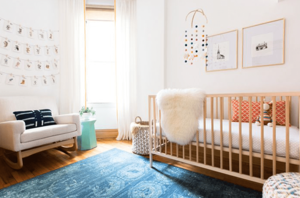 Baby girl nursery with wood crib and flooring, blue carpet and white rocking chair inspired by Modern design.