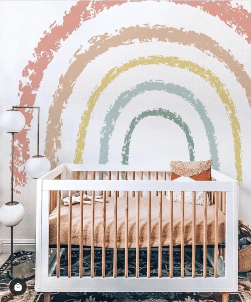Baby girl nursery with minimalist white crib, soft pastel mattress, white floor lamps and a large wall decal that shows a rainbow in muted colors.