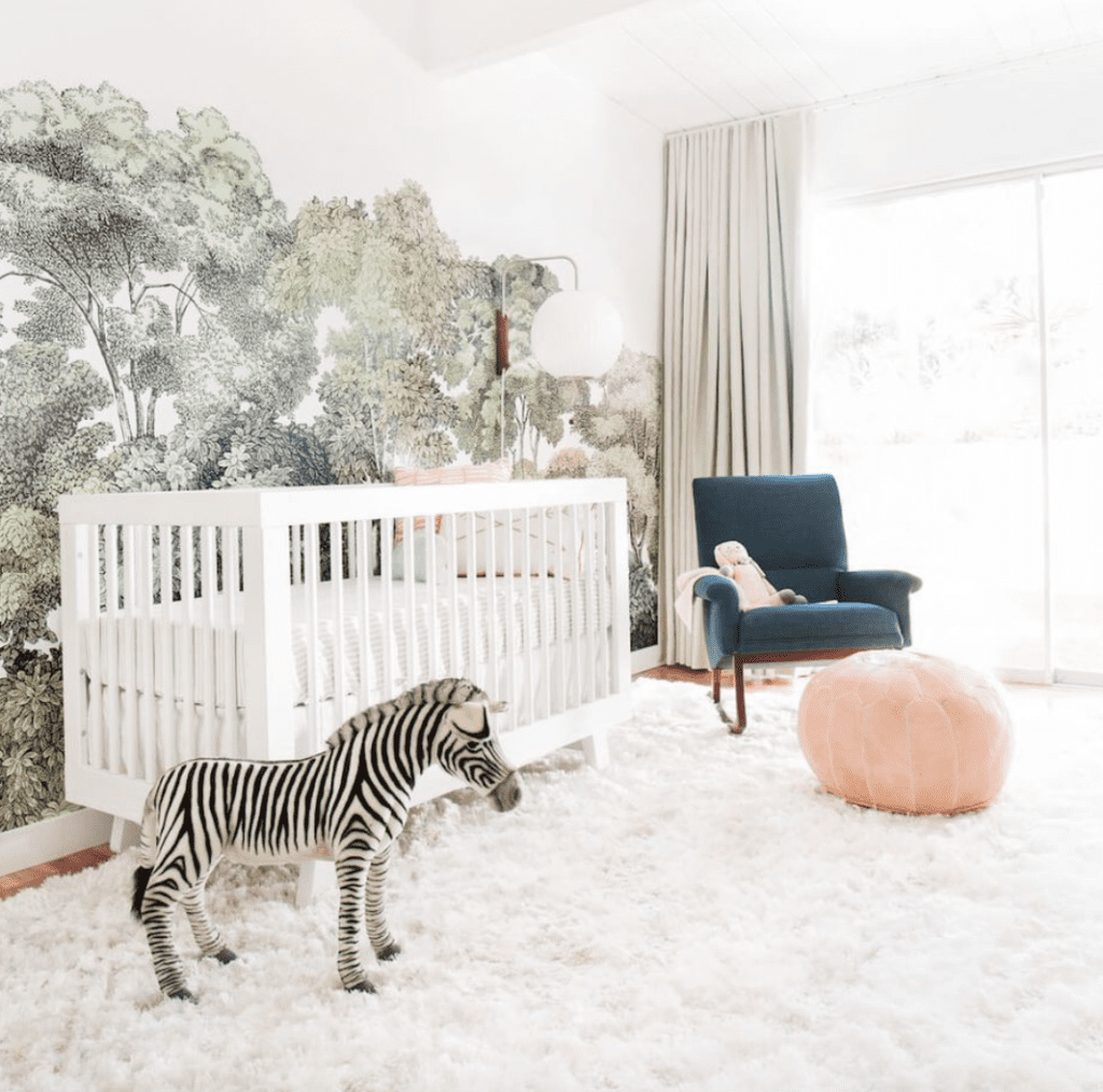 Baby girl nursery with gray wall to ceiling curtains, white carpet, coral pouf, blue gray couch, white crib and accent wall inspiored by tropical jungle.