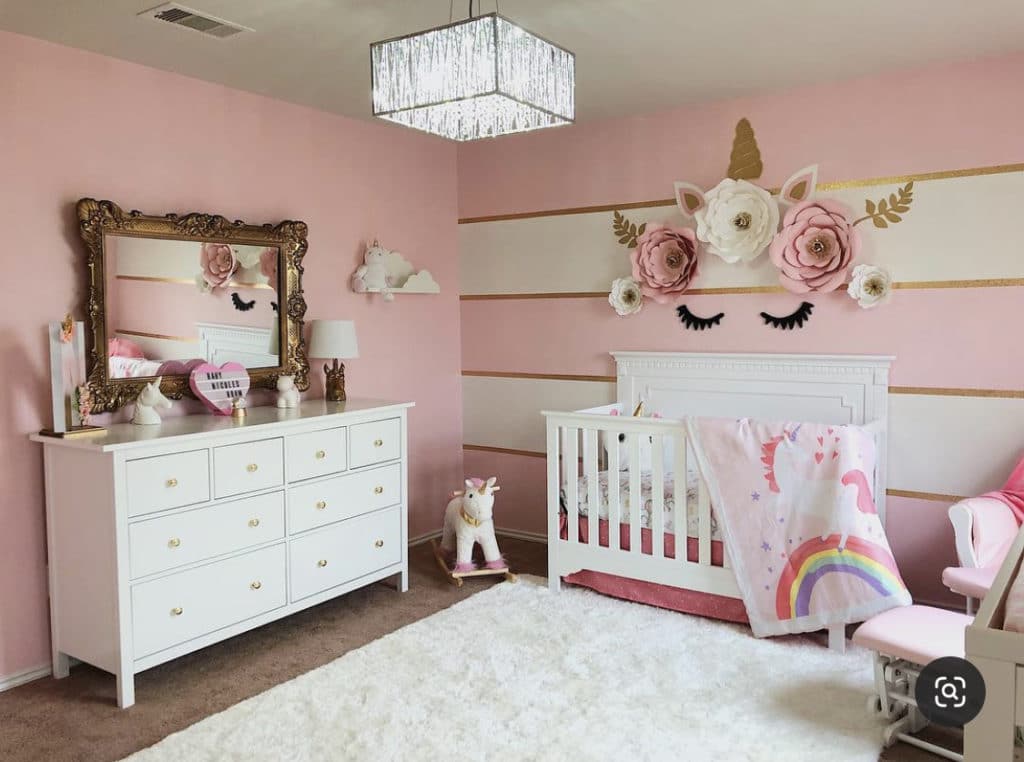 Baby girl nursery with blush walls,  gold, pink, and white tones, floral wall decor, a plush rug, and white chest of drawers representing a unicorn themed space.