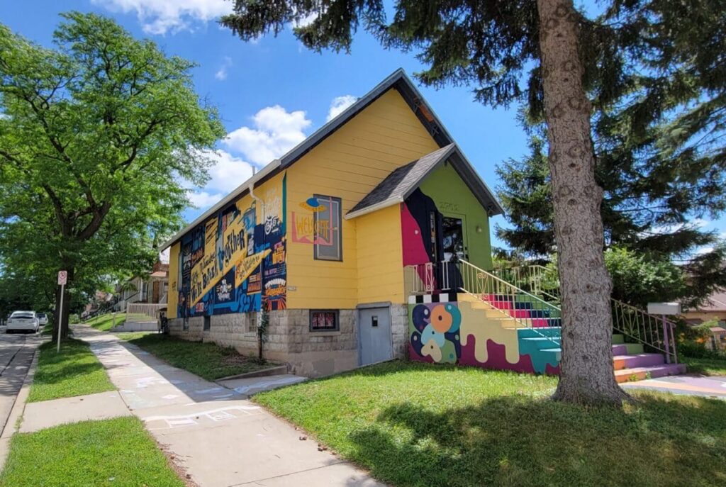 Colorful exterior of Bay View Printing Co. in Milwaukee, featuring a community mural, painted steps, and bright yellow siding on a sunny day.
