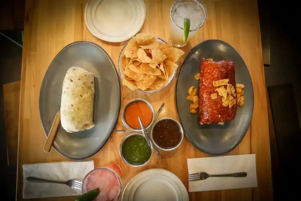 A top view of 2 burritos with three kinds of sauces and a bowl of chips