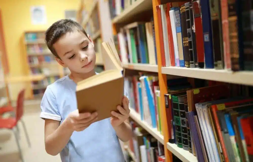 Young boy reading a book in a library aisle.