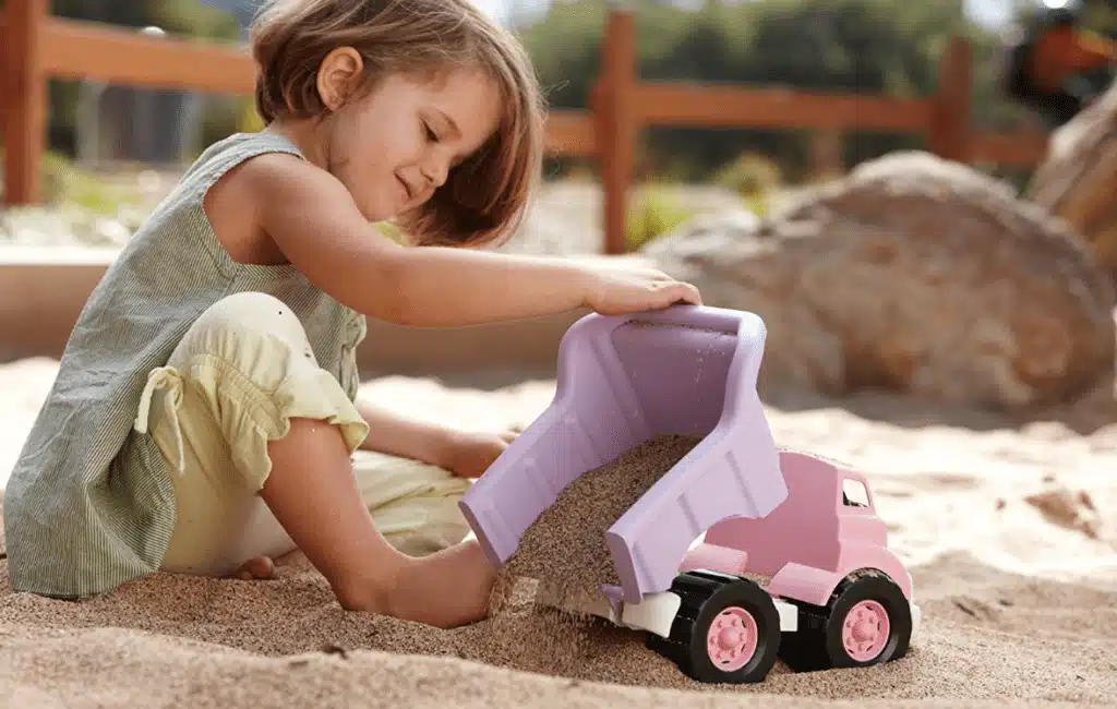 A little girl playing with a purple dump truck toy in a sand box