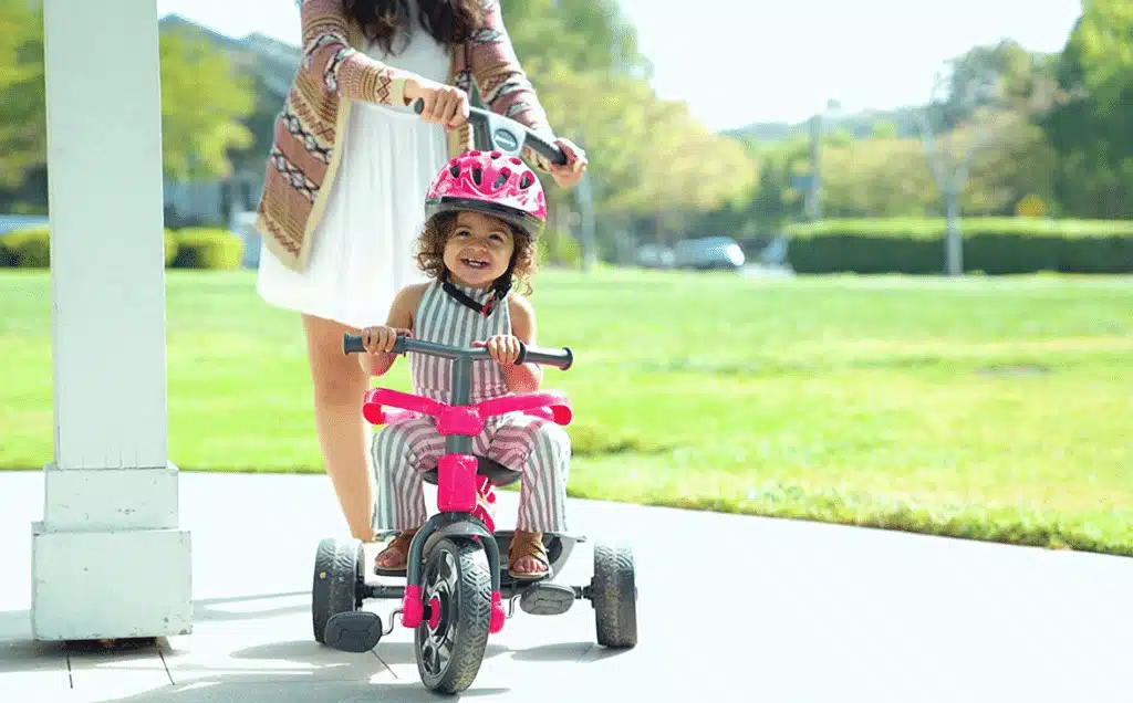 A little girl with head gear riding on a bikewhile being pushed by her mom in the park