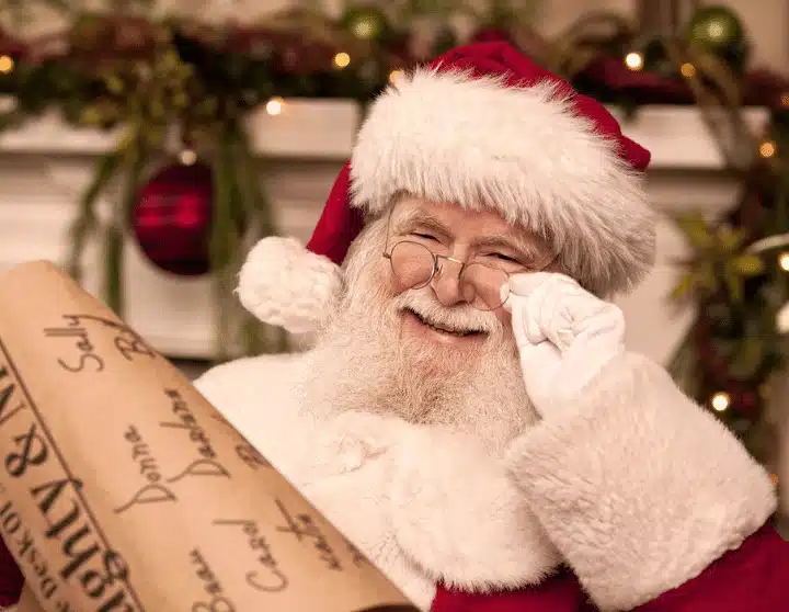 A smiling Santa holding his spectacles on one hand and the nauight and lice list on another