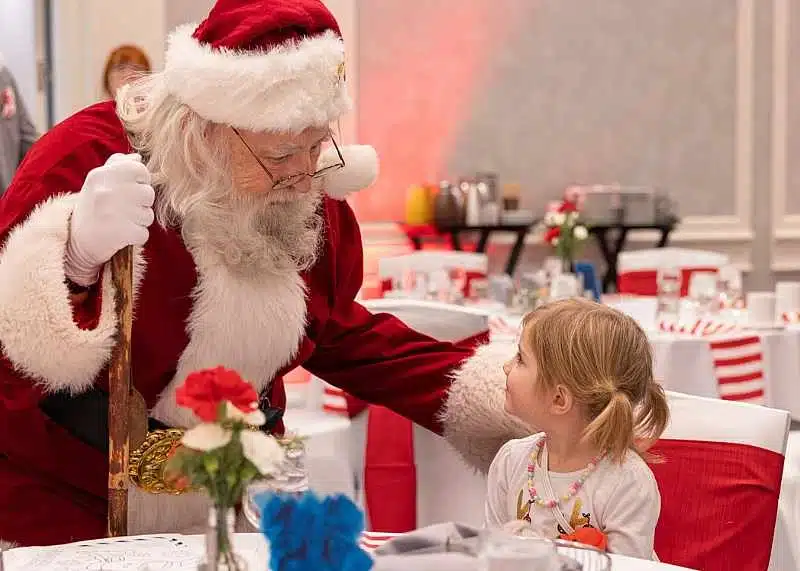 Santa holding a wooden can and talking to a sitting girl in pigtails and white graphic shirt.